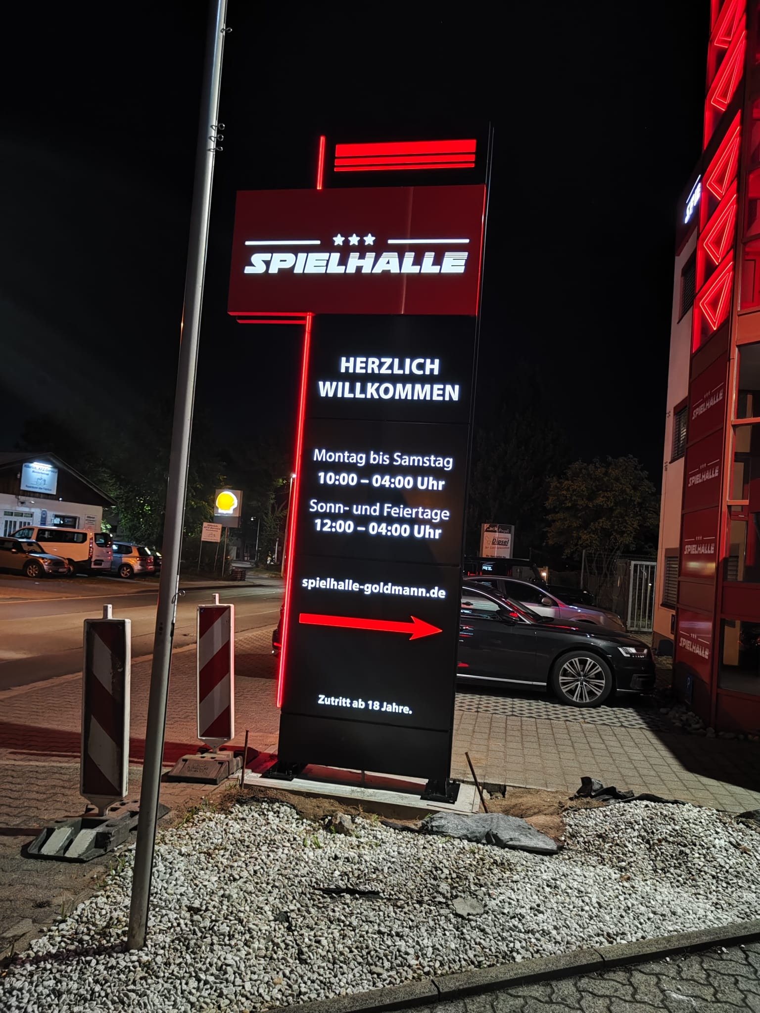 Podświetlony pylon reklamowy z nazwą 'Spielhalle', godzinami otwarcia i adresem strony internetowej, ustawiony przed budynkiem z podobnym neonowym oznakowaniem.