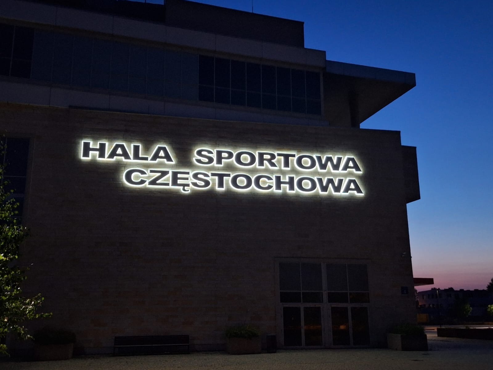 Elewacja hali sportowej w Częstochowie z podświetlanymi literami tworzącymi napis. Widok z dołu na budynek o zmierzchu.