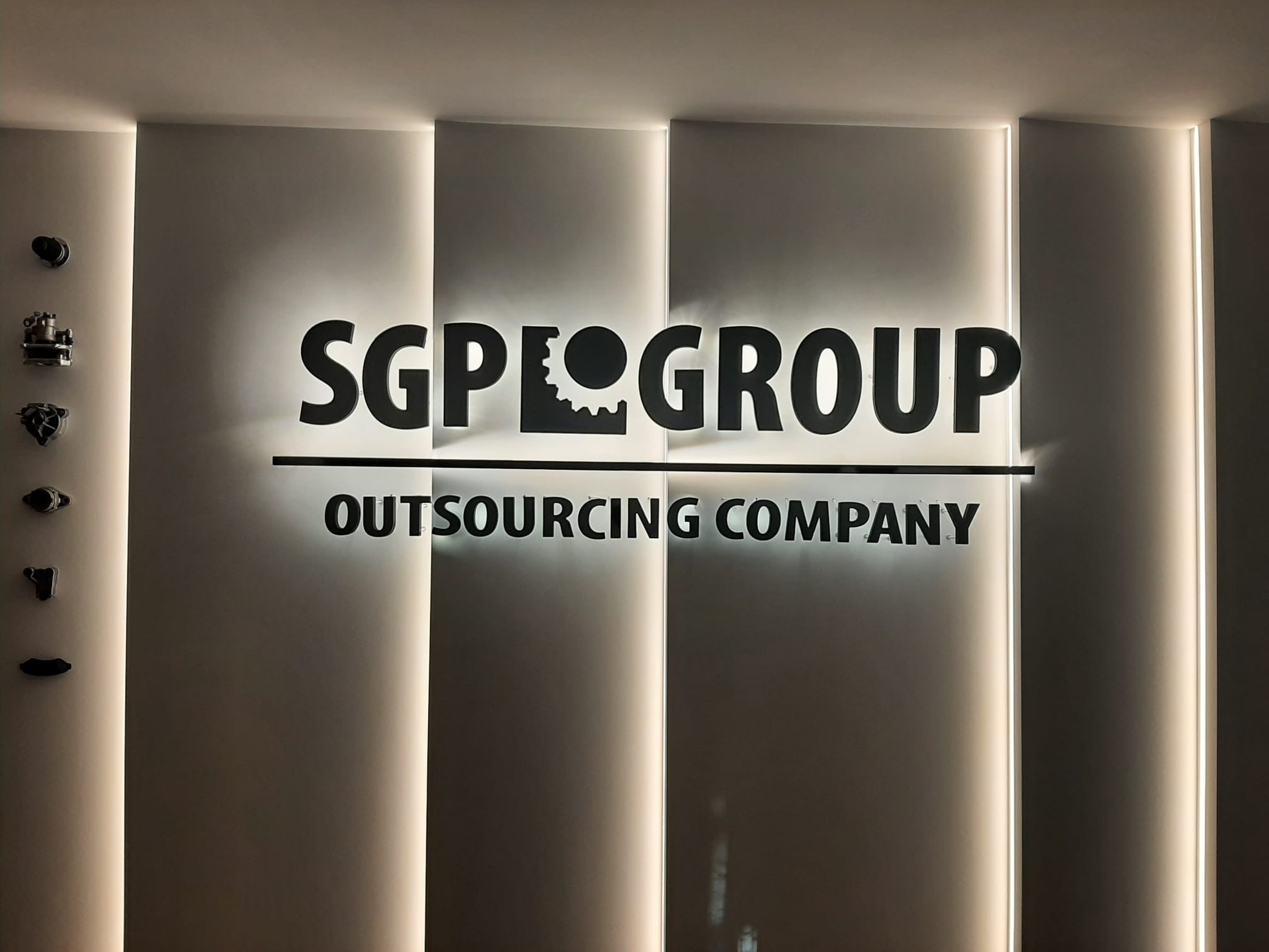Świecący napis SGP GROUP Outsourcing Company na ścianie, podświetlony od tyłu, obok zamontowane elementy metalowe, pionowe linie podświetlenia po bokach.