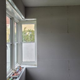Janusz Bąk - Ściana z płyt gipsowo-kartonowych w trakcie montażu, widoczne łączenia i wkręty. Okno z widokiem na zieleń. Narzędzia na parapecie.