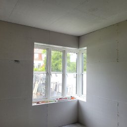 Janusz Bąk - Pokój w trakcie montażu płyt gipsowo-kartonowych, widoczne łączenia i wkręty. Okno narożne z widokiem na zewnątrz. Etap przygotowania do dalszych prac wykończeniowych.