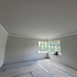 Janusz Bąk - Pokój po szpachlowaniu, przygotowany do malowania. Widoczne listwy przysufitowe, okno z widokiem na zieleń i zabezpieczona podłoga. Ściany gładkie, w jasnym odcieniu szarości.