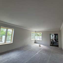 Janusz Bąk - Wnętrze po szpachlowaniu i malowaniu na szaro, z zabezpieczoną podłogą, widoczne okna i sztukateria. Przygotowanie do dalszych prac wykończeniowych.