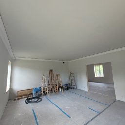 Janusz Bąk - Jasne wnętrze po remoncie z listwami dekoracyjnymi, drabiny malarskie i odkurzacz przemysłowy. Podłoga zabezpieczona taśmą. Widok na sąsiednie pomieszczenie.