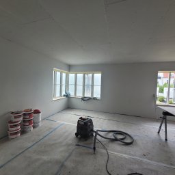 Janusz Bąk - Pokój w trakcie prac wykończeniowych: ściany po szpachlowaniu, zabezpieczona podłoga, widoczne puszki z farbą i odkurzacz przemysłowy. Okna z widokiem na zieleń.