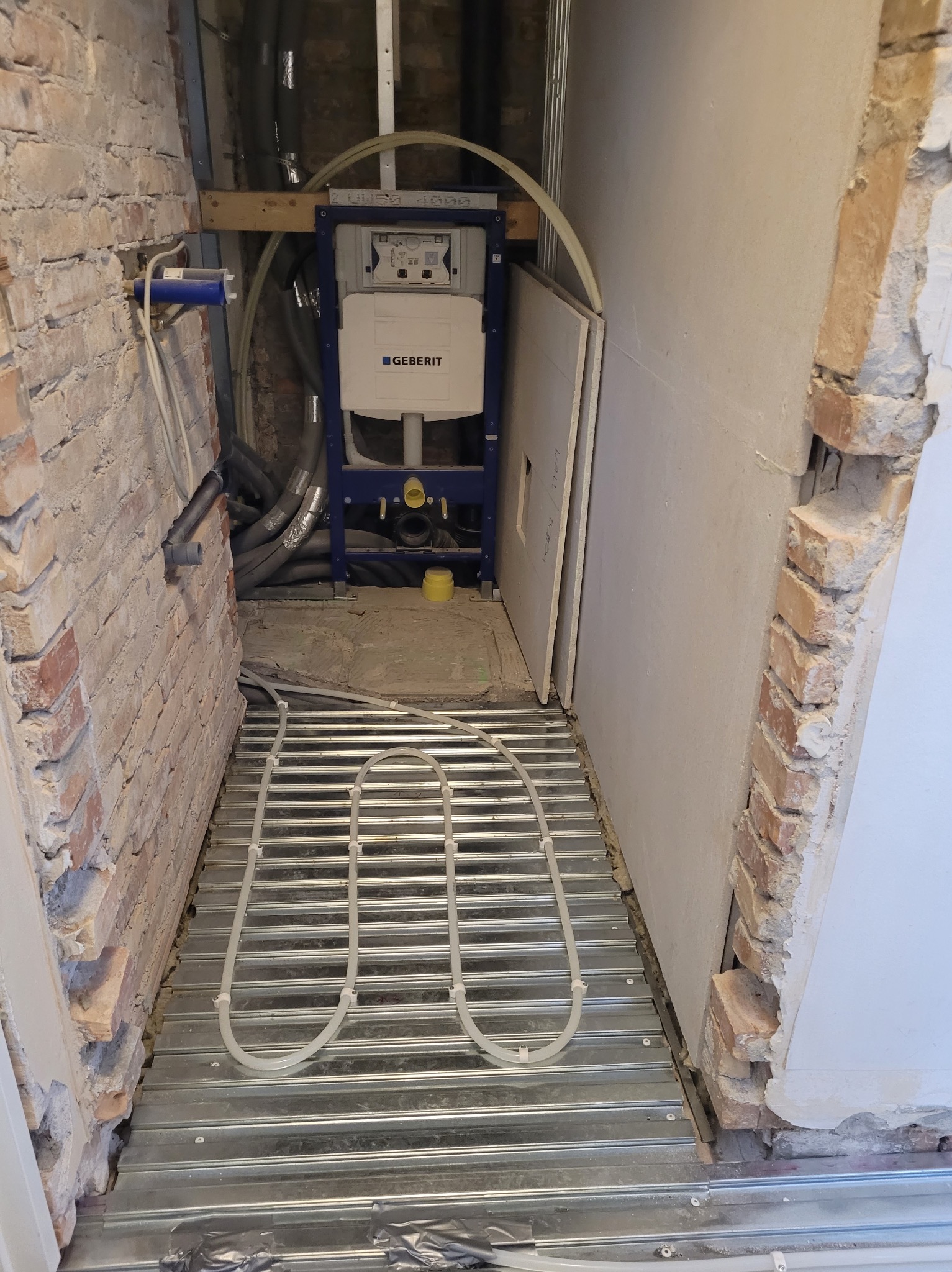 Remont łazienki w trakcie: widoczna instalacja ogrzewania podłogowego na metalowej konstrukcji, stelaż podtynkowy Geberit i fragmenty ścian z cegły i płyt karton-gips.