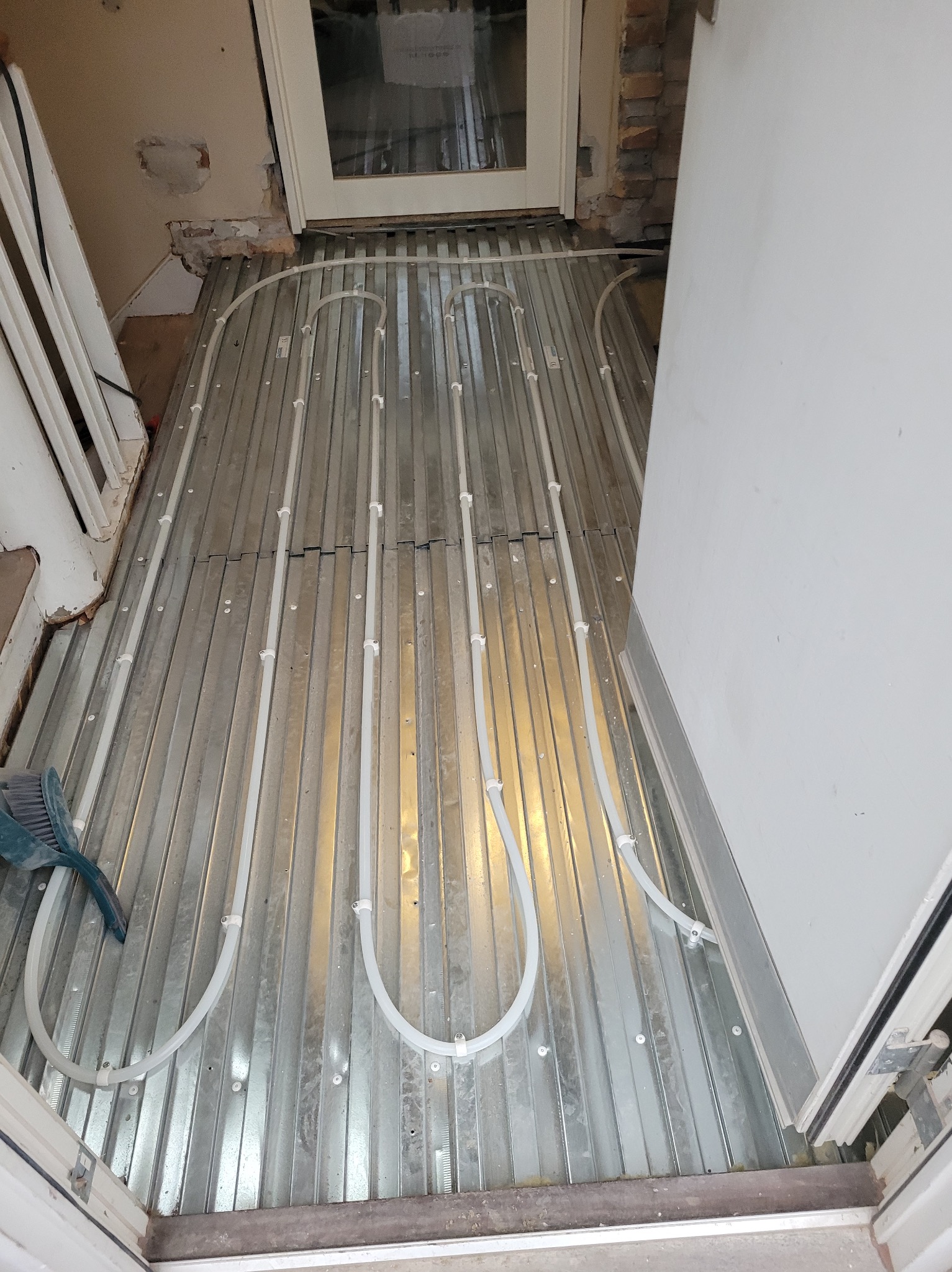 Montaż ogrzewania podłogowego: białe rurki na metalowej płycie, widoczne elementy konstrukcyjne podłoża i fragment ściany z ubytkiem tynku. Etap prac przed wylaniem posadzki.