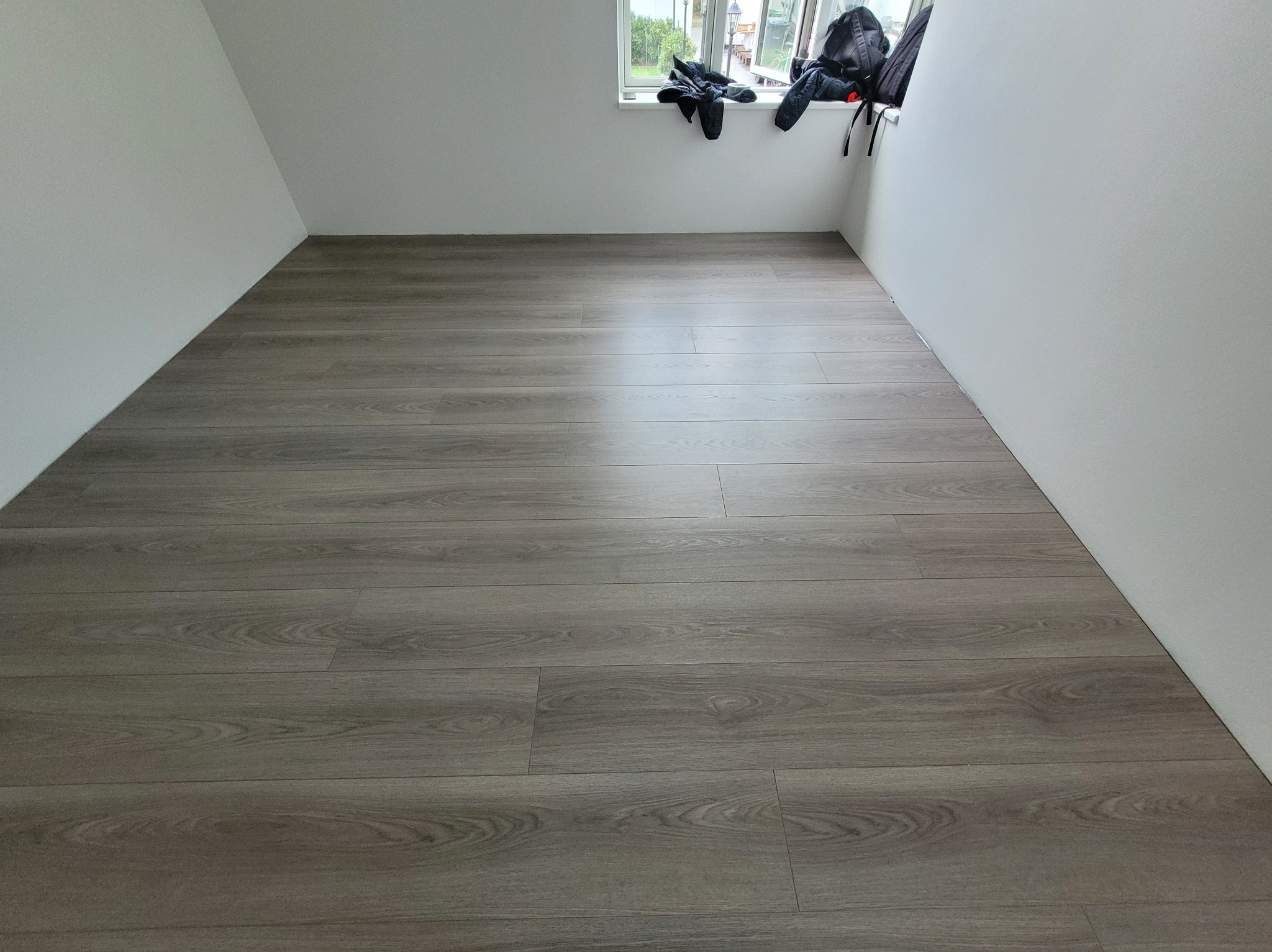Ułożone panele podłogowe w pokoju ze skośnymi ścianami i oknem, na parapecie leżą ubrania i plecak. Widok na szare panele z bliska.