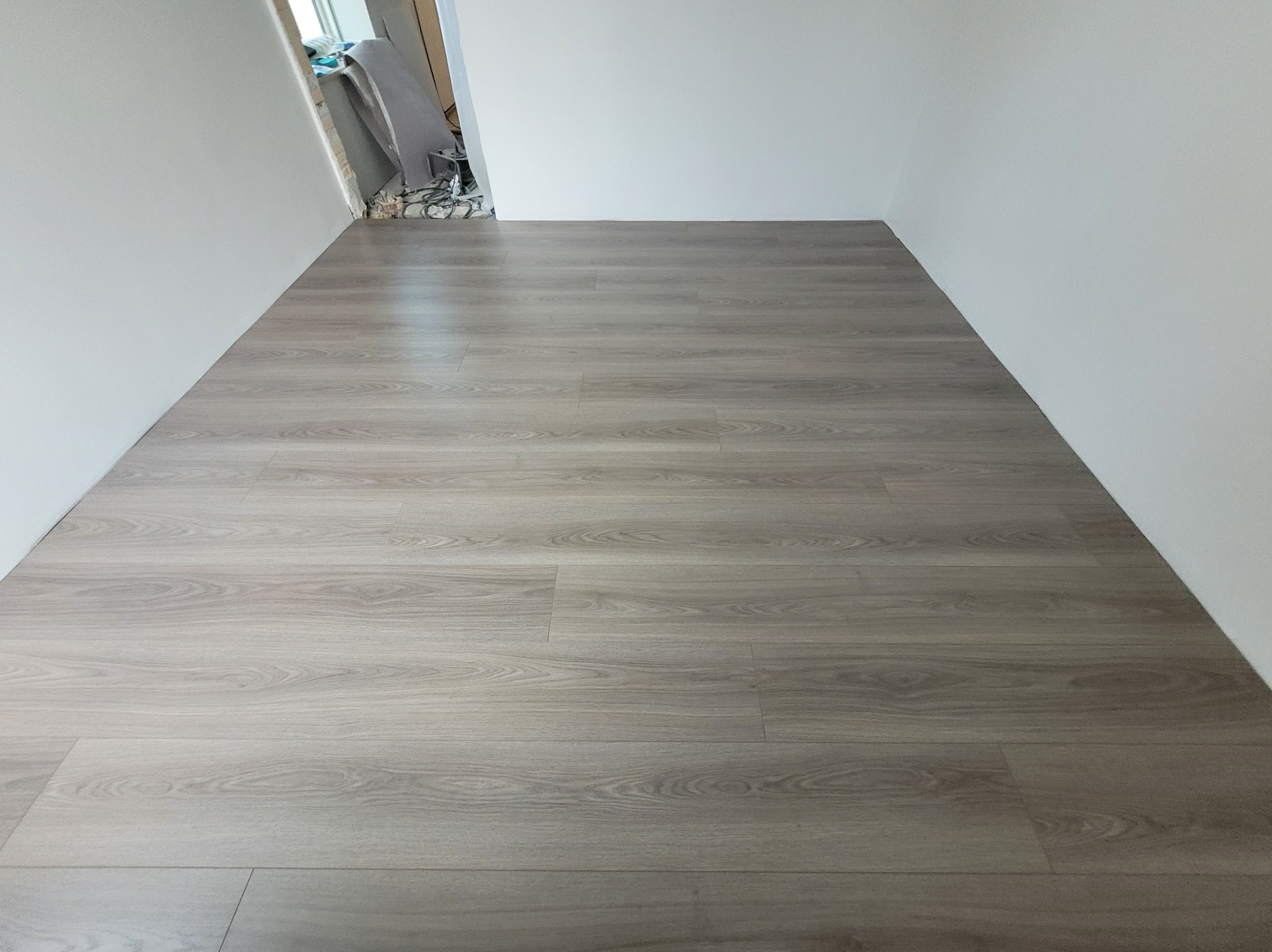 Ułożone panele podłogowe w wąskim korytarzu, widok z góry. Szare panele z wzorem drewna, białe ściany. W tle widoczne kable i materiały budowlane.