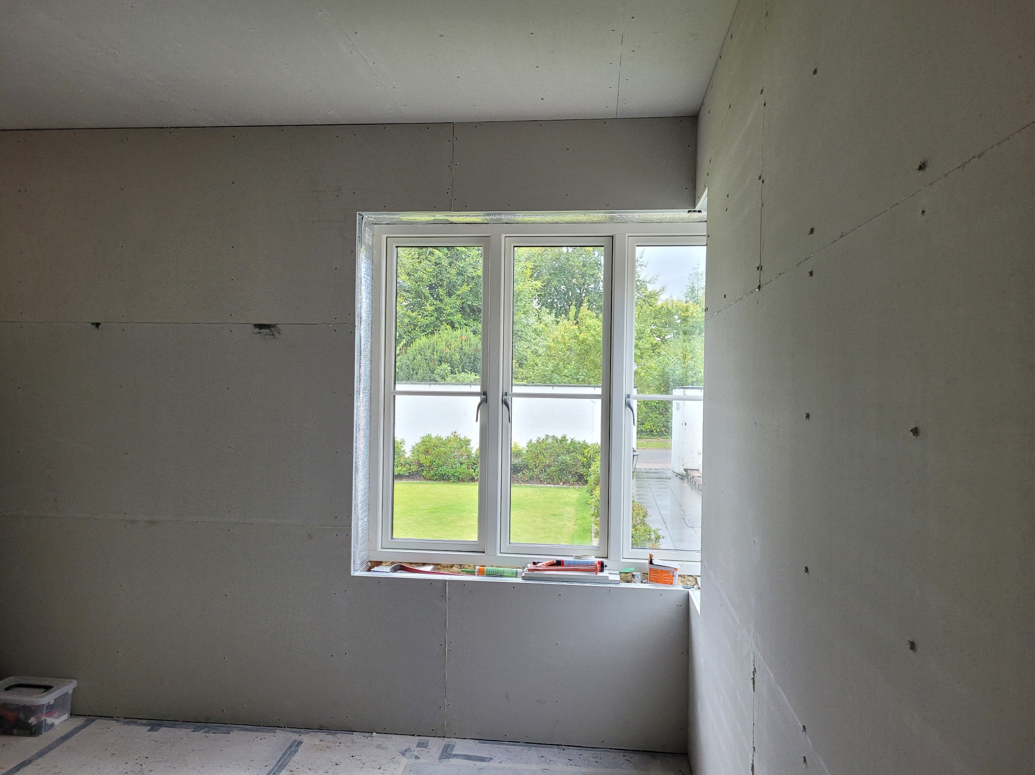 Pokój w trakcie prac wykończeniowych: ściany i sufit pokryte płytami gipsowo-kartonowymi, okno z widokiem na zieleń. Widoczne narzędzia na parapecie.
