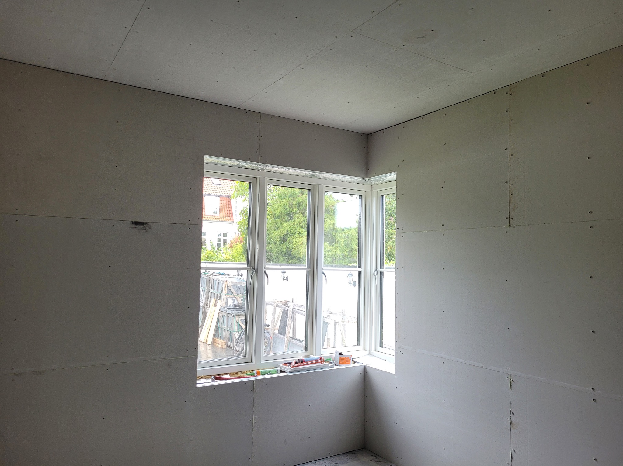 Pokój w trakcie montażu płyt gipsowo-kartonowych, widoczne łączenia i wkręty. Okno narożne z widokiem na zewnątrz. Etap przygotowania do dalszych prac wykończeniowych.