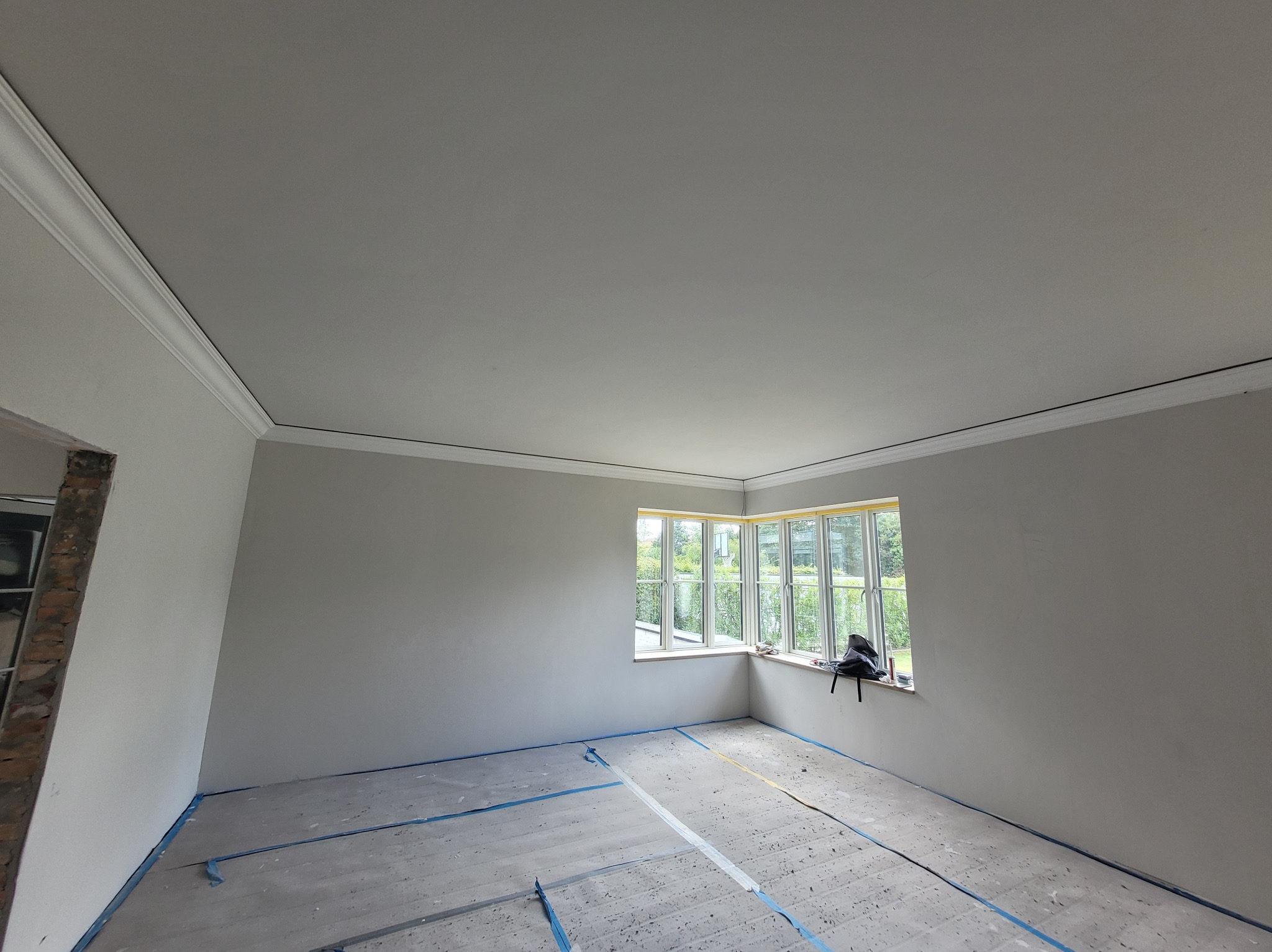 Pokój po szpachlowaniu, przygotowany do malowania. Widoczne listwy przysufitowe, okno z widokiem na zieleń i zabezpieczona podłoga. Ściany gładkie, w jasnym odcieniu szarości.