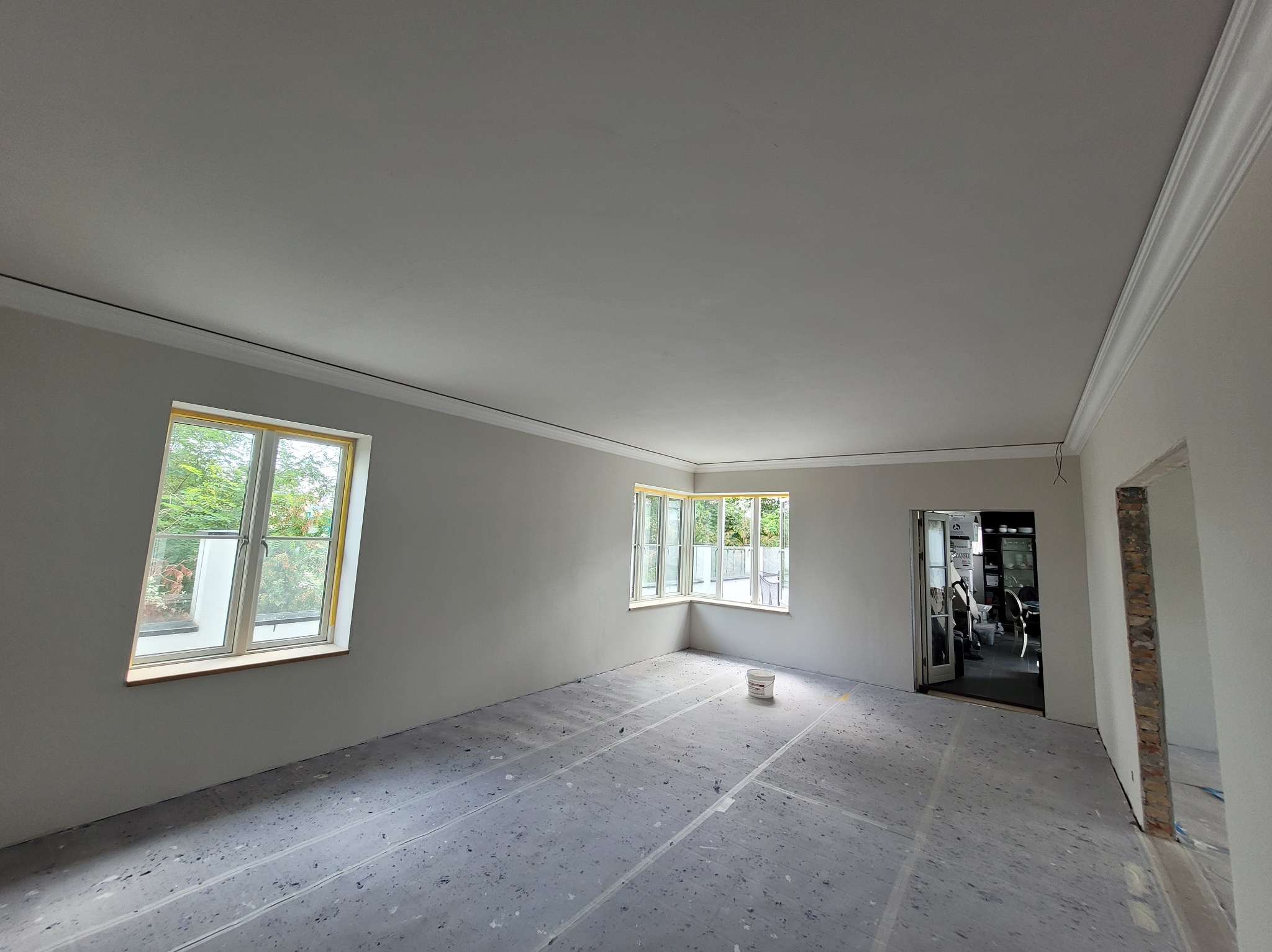 Wnętrze po szpachlowaniu i malowaniu na szaro, z zabezpieczoną podłogą, widoczne okna i sztukateria. Przygotowanie do dalszych prac wykończeniowych.