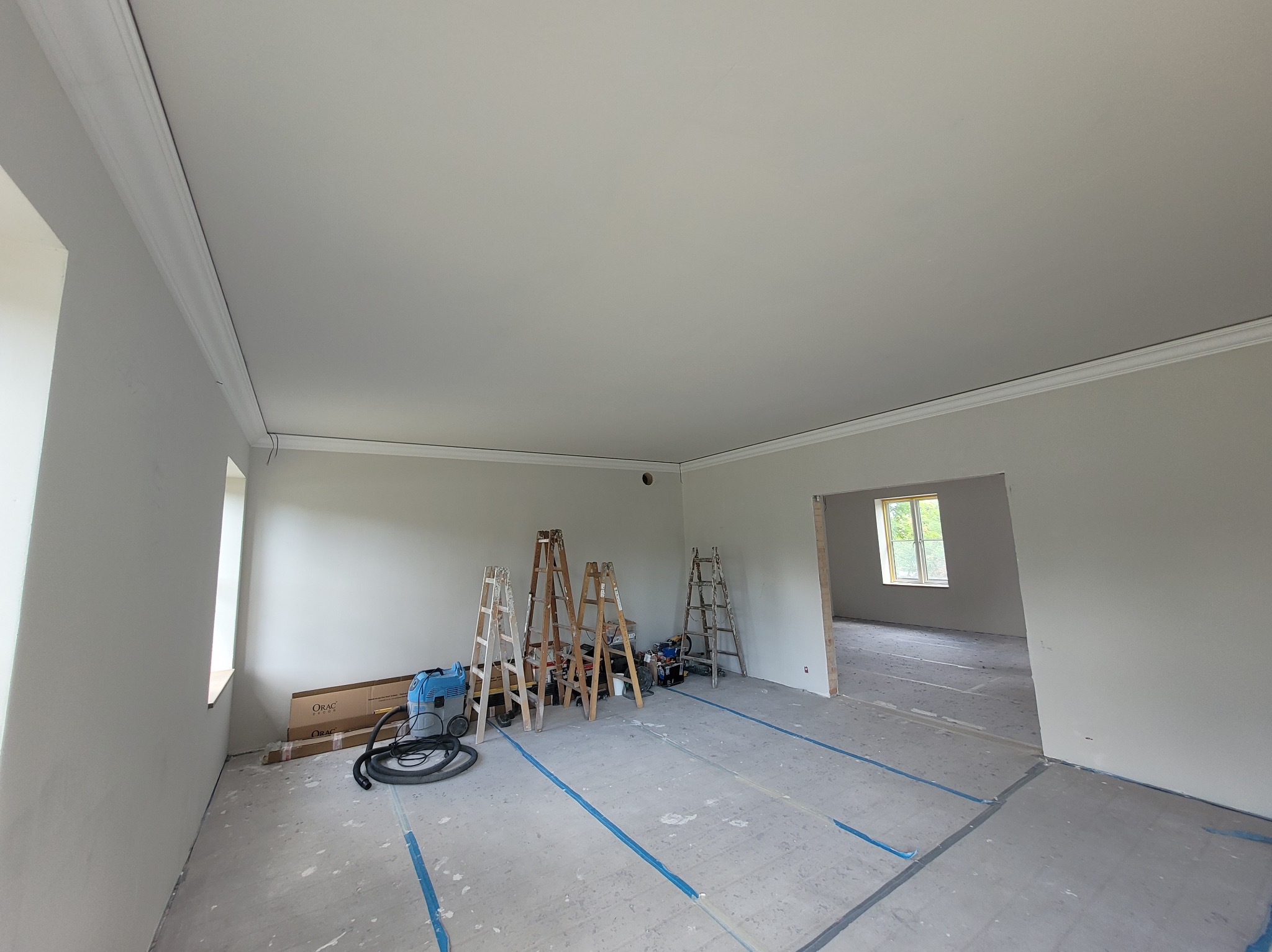 Jasne wnętrze po remoncie z listwami dekoracyjnymi, drabiny malarskie i odkurzacz przemysłowy. Podłoga zabezpieczona taśmą. Widok na sąsiednie pomieszczenie.