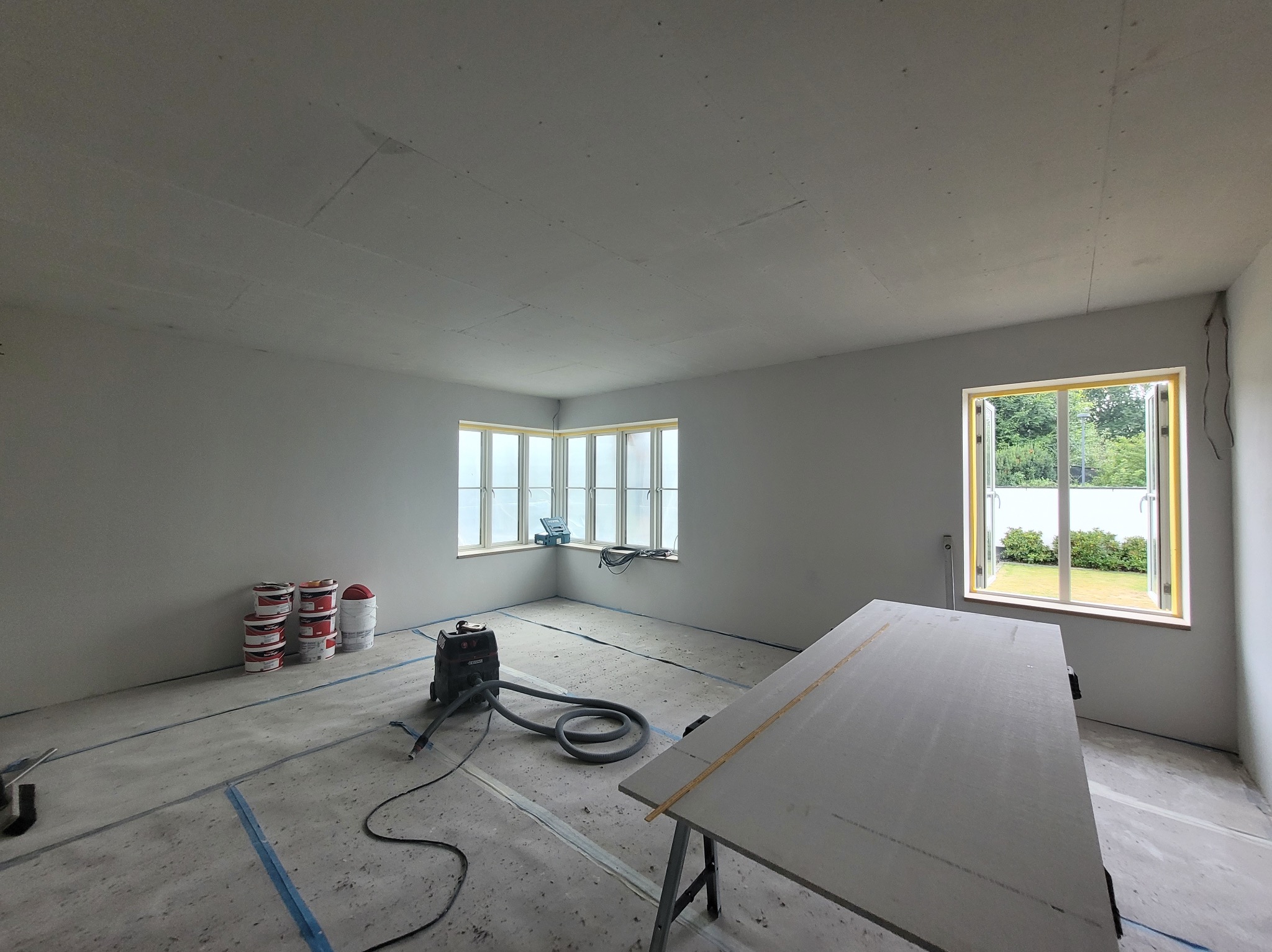 Przygotowane pomieszczenie do prac wykończeniowych: zabezpieczona podłoga, ustawiony stół roboczy, widoczne farby i odkurzacz przemysłowy. Dwa okna z widokiem na zieleń.