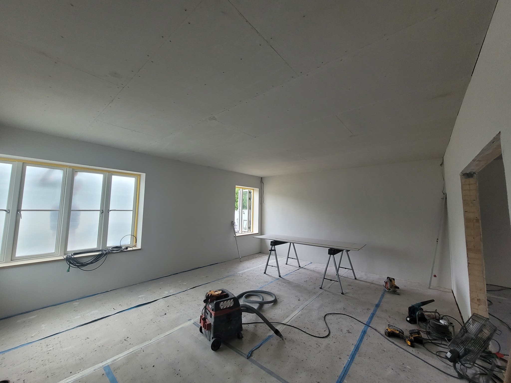 Pokój w trakcie prac wykończeniowych: zamontowane płyty na suficie, przygotowane narzędzia, odkurzacz przemysłowy. Widoczne okna i surowe wykończenie ścian.