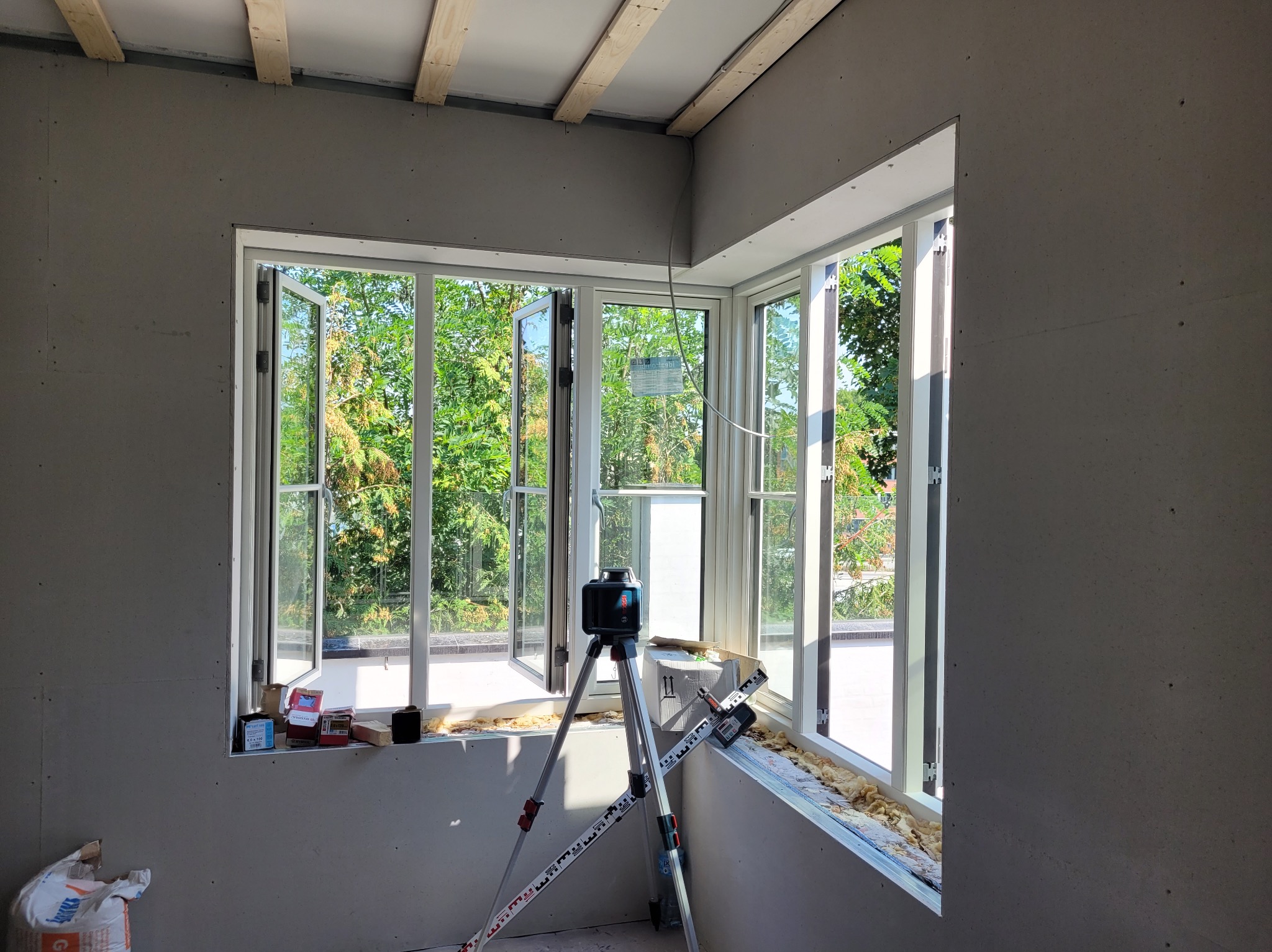 Wnętrze w trakcie prac z płytami gipsowo-kartonowymi, widoczny laser krzyżowy na statywie, okno z widokiem na zieleń. Ściany przygotowane do dalszych prac wykończeniowych.