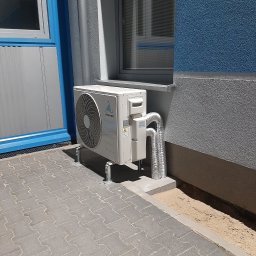 FP - SYSTEM FILIP PÓŁTORAK - Biała jednostka zewnętrzna klimatyzatora marki Vesser zamontowana na wspornikach przy ścianie budynku z niebieskim i szarym tynkiem, z widocznymi elastycznymi przewodami odprowadzającymi.