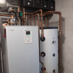 FP - SYSTEM FILIP PÓŁTORAK - Szara pompa ciepła marki Nibe BA-SVM 10-200 z miedzianymi rurami i czarnymi elementami sterującymi na tle szarej ściany z płytek.