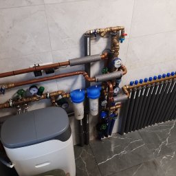 FP - SYSTEM FILIP PÓŁTORAK - Węzeł cieplny z rozdzielaczem, filtrami wody, zmiękczaczem i licznikiem ciepła, rury miedziane z izolacją, szare płytki na ścianie i podłodze.