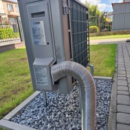 FP - SYSTEM FILIP PÓŁTORAK - Szara jednostka zewnętrzna z logo NIBE, zamontowana na żwirowym podłożu z obrzeżem, z widoczną giętką, metalową rurą odprowadzającą. W tle trawnik, chodnik z kostki i ogrodzenie.