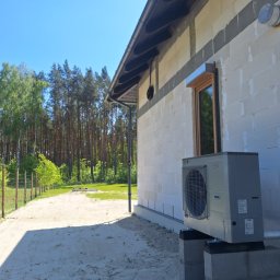 FP - SYSTEM FILIP PÓŁTORAK - Zamontowana pompa ciepła przy ścianie domu z pustaków, widok z zewnątrz w słoneczny dzień. Elementy instalacji widoczne w tle, las w oddali.