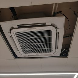 FP - SYSTEM FILIP PÓŁTORAK - Kasetonowa klimatyzacja sufitowa wkomponowana w podwieszany sufit, widok od dołu. Widoczne elementy instalacji i przewody. Oświetlenie liniowe LED w tle.