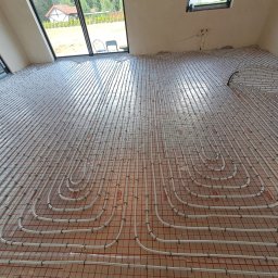 FP - SYSTEM FILIP PÓŁTORAK - Ułożone rury ogrzewania podłogowego na folii izolacyjnej z widocznymi oknami i fragmentem domu na zewnątrz. Nowoczesna instalacja w jasnym pomieszczeniu.