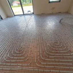 Ułożone rury ogrzewania podłogowego na folii izolacyjnej z widocznymi oknami i fragmentem domu na zewnątrz. Nowoczesna instalacja w jasnym pomieszczeniu.