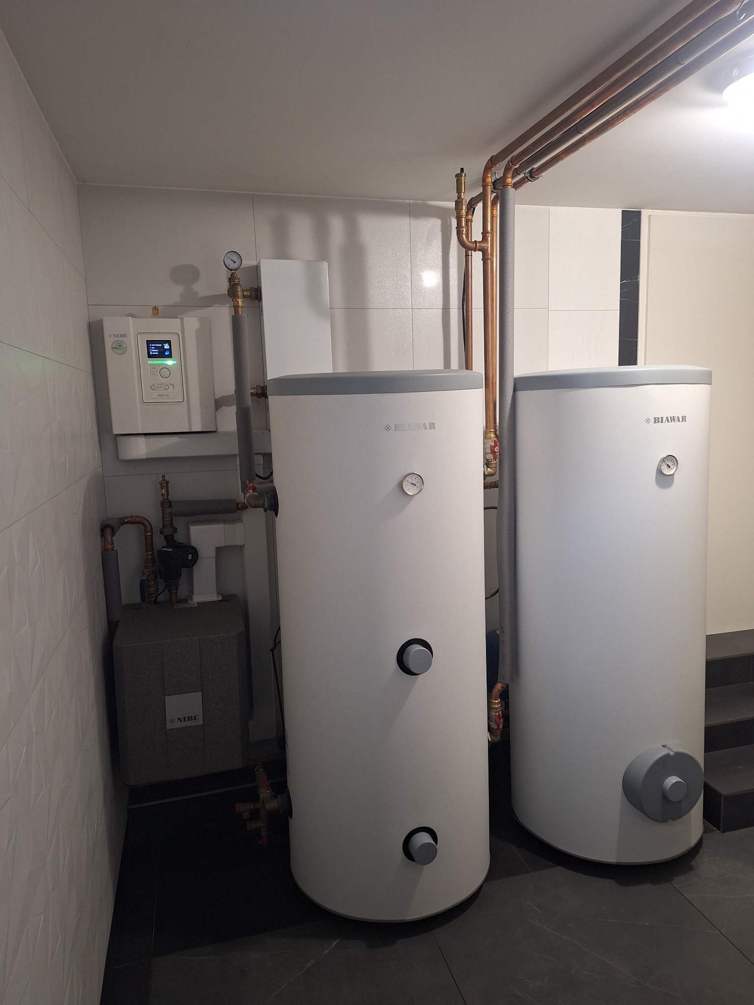 Dwa białe zbiorniki akumulacyjne Biawar z manometrami w pomieszczeniu z płytkami, widoczne rury miedziane, sterownik Nibe SMO 20 i szary blok Nibe.