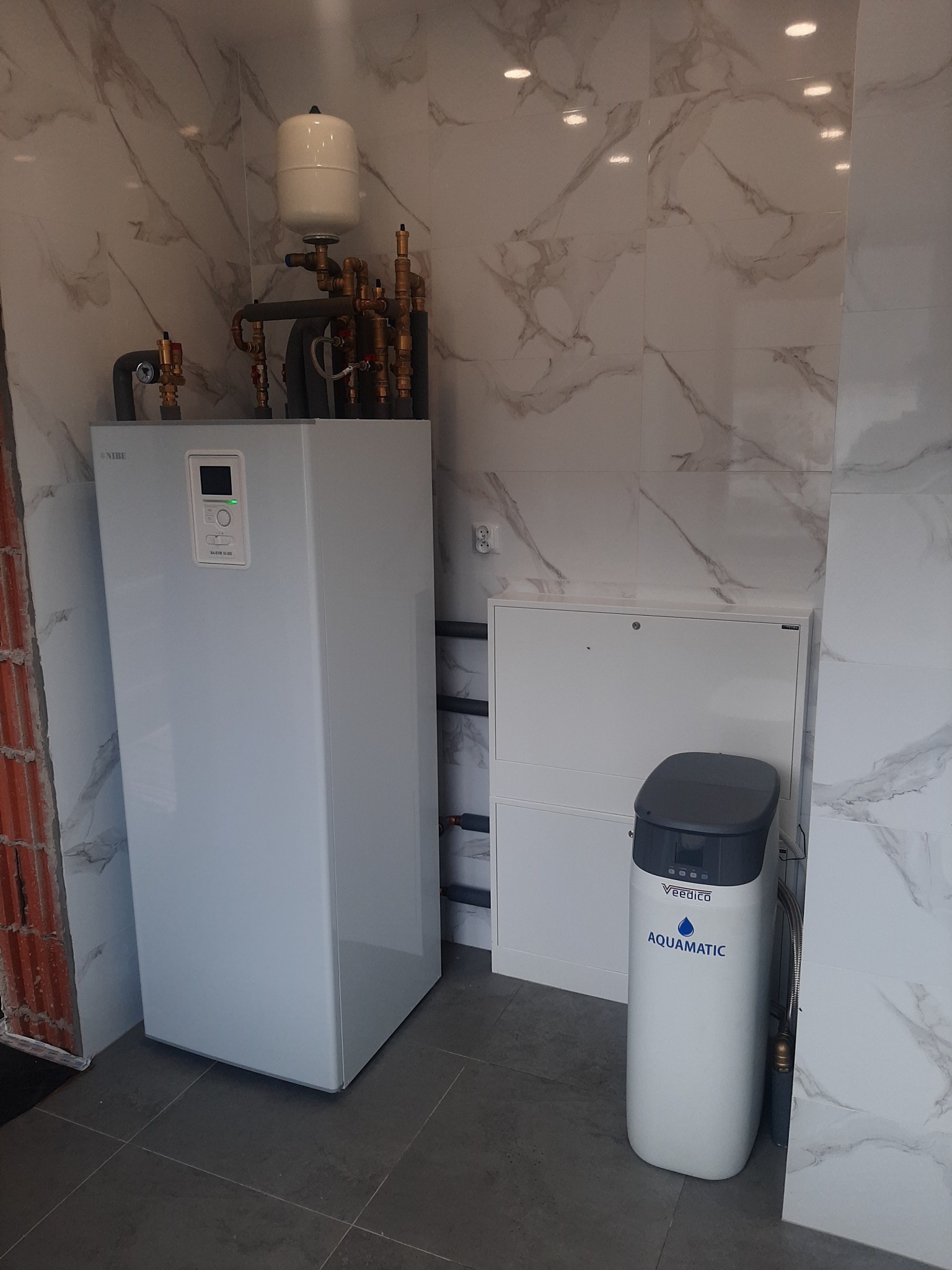 Biała instalacja grzewcza Nibe z zbiornikiem wyrównawczym i filtrem wody Aquamatic na tle marmurowych płytek i surowej ściany z cegły.