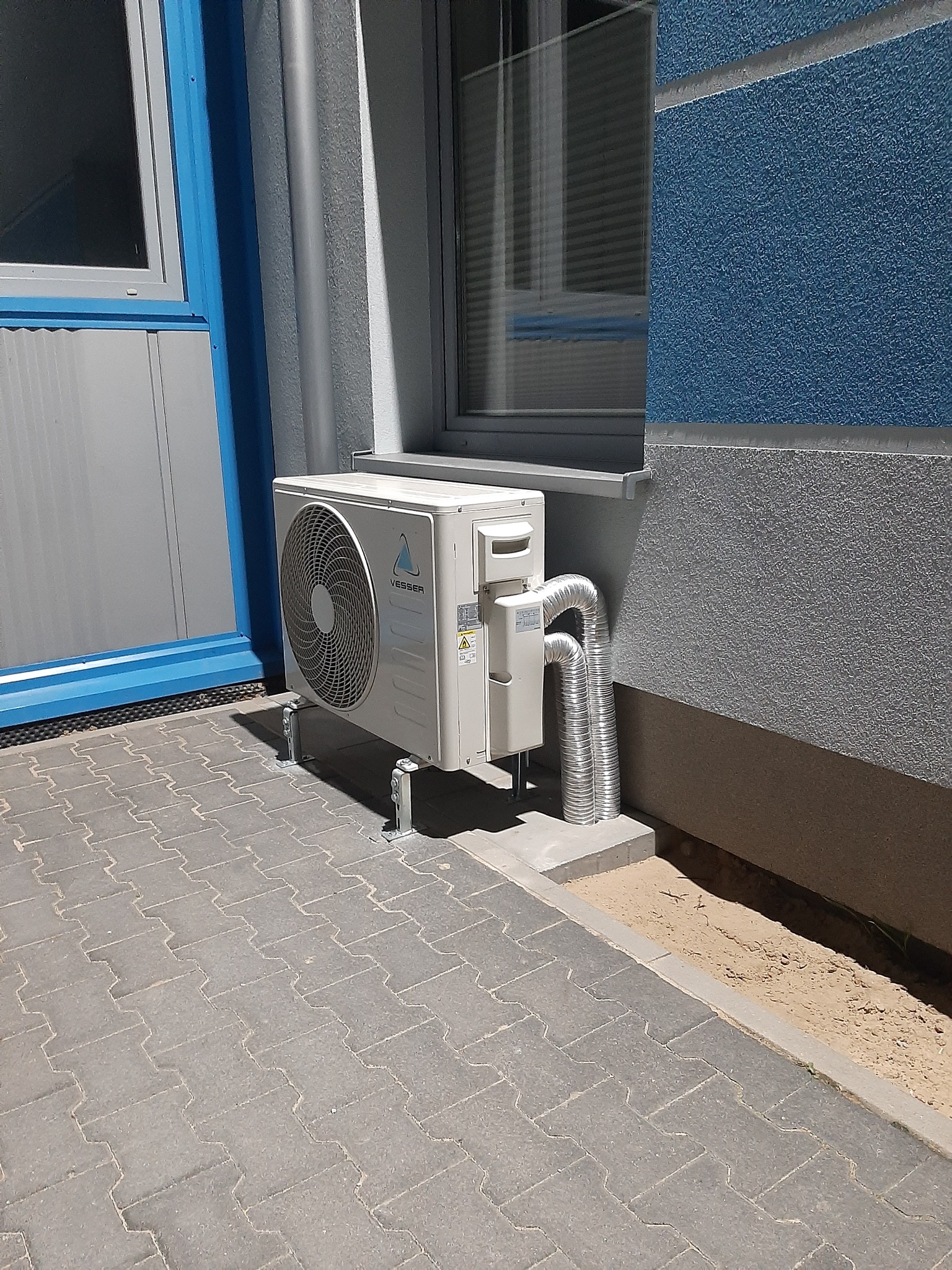 Biała jednostka zewnętrzna klimatyzatora marki Vesser zamontowana na wspornikach przy ścianie budynku z niebieskim i szarym tynkiem, z widocznymi elastycznymi przewodami odprowadzającymi.