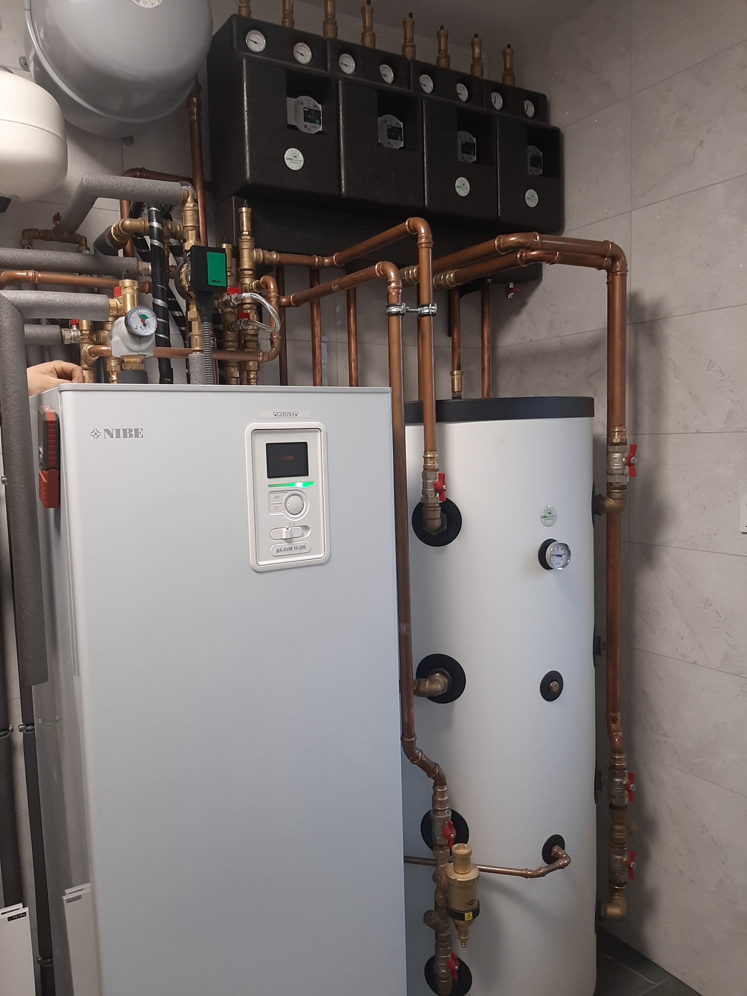 Szara pompa ciepła marki Nibe BA-SVM 10-200 z miedzianymi rurami i czarnymi elementami sterującymi na tle szarej ściany z płytek.