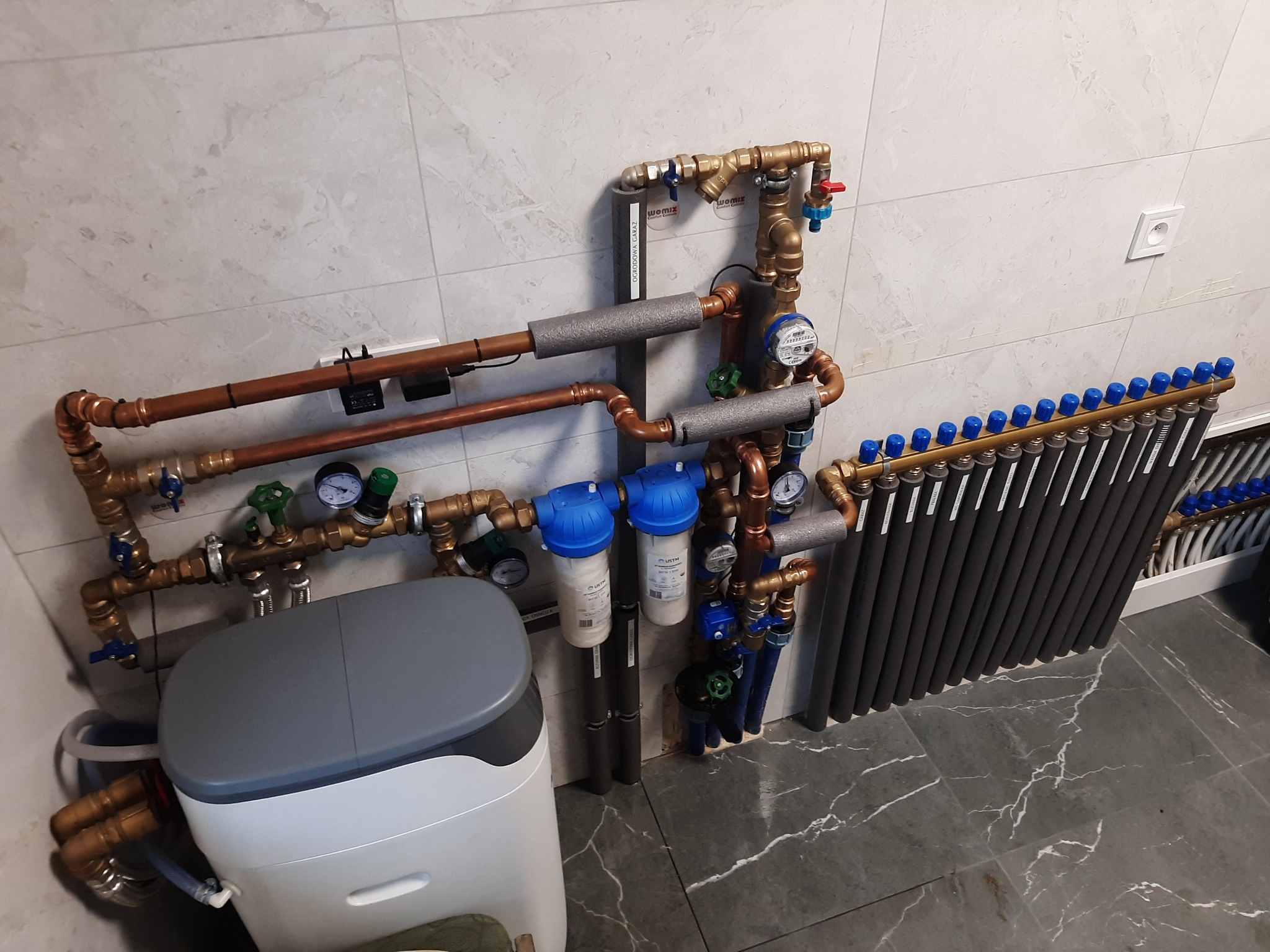 Węzeł cieplny z rozdzielaczem, filtrami wody, zmiękczaczem i licznikiem ciepła, rury miedziane z izolacją, szare płytki na ścianie i podłodze.