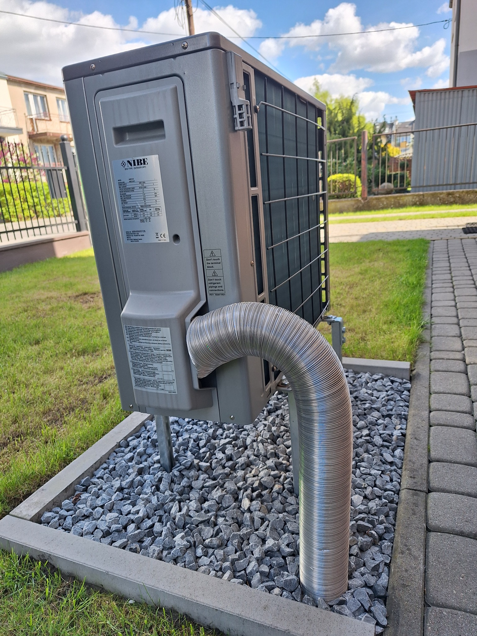 Szara jednostka zewnętrzna z logo NIBE, zamontowana na żwirowym podłożu z obrzeżem, z widoczną giętką, metalową rurą odprowadzającą. W tle trawnik, chodnik z kostki i ogrodzenie.