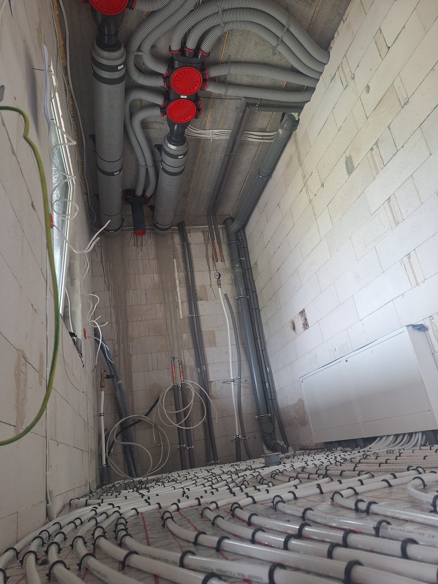 Rozłożone rury ogrzewania podłogowego na siatce, widoczne rury wentylacyjne pod sufitem i rury instalacji hydraulicznej na ścianie w surowym stanie.