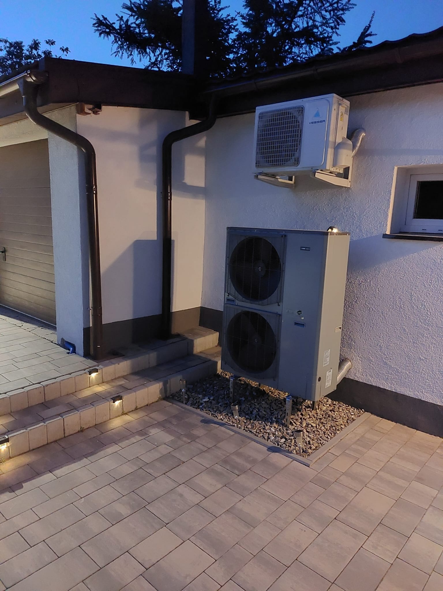 Zewnętrzna jednostka klimatyzacji i pompa ciepła zamontowane na elewacji budynku z oświetlonymi schodami wejściowymi i widoczną rynną spustową.