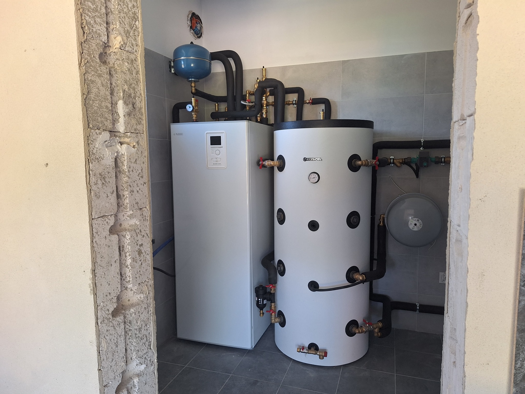 Biały piec CO z zbiornikiem Kospel i niebieskim naczyniem przeponowym, instalacja z czarnych rur, szare płytki na ścianie, widok zza surowej framugi.