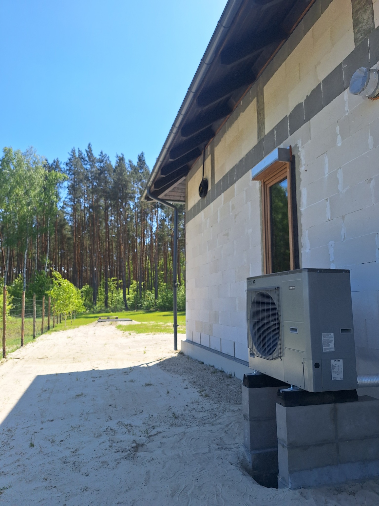 Zamontowana pompa ciepła przy ścianie domu z pustaków, widok z zewnątrz w słoneczny dzień. Elementy instalacji widoczne w tle, las w oddali.