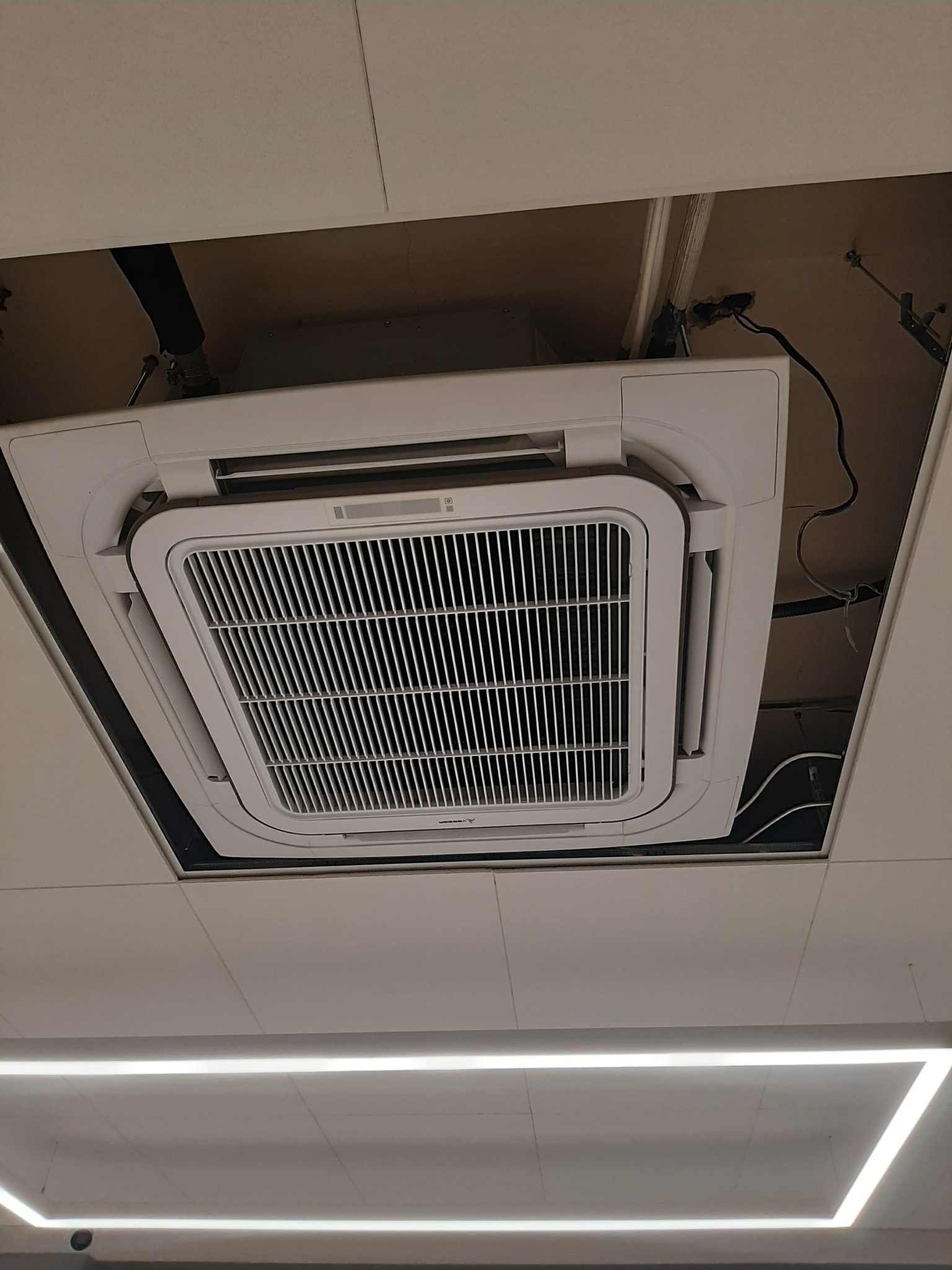Kasetonowa klimatyzacja sufitowa wkomponowana w podwieszany sufit, widok od dołu. Widoczne elementy instalacji i przewody. Oświetlenie liniowe LED w tle.