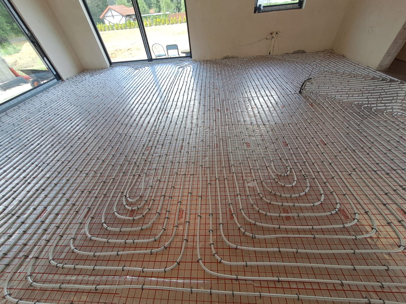 Ułożone rury ogrzewania podłogowego na folii izolacyjnej z widocznymi oknami i fragmentem domu na zewnątrz. Nowoczesna instalacja w jasnym pomieszczeniu.