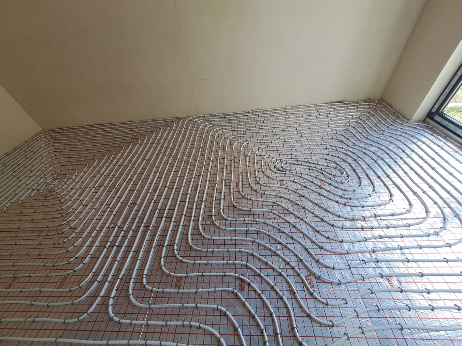 Białe rury ogrzewania podłogowego ułożone spiralnie na folii izolacyjnej z czerwoną siatką w jasnym wnętrzu, widoczne okno z prawej strony.
