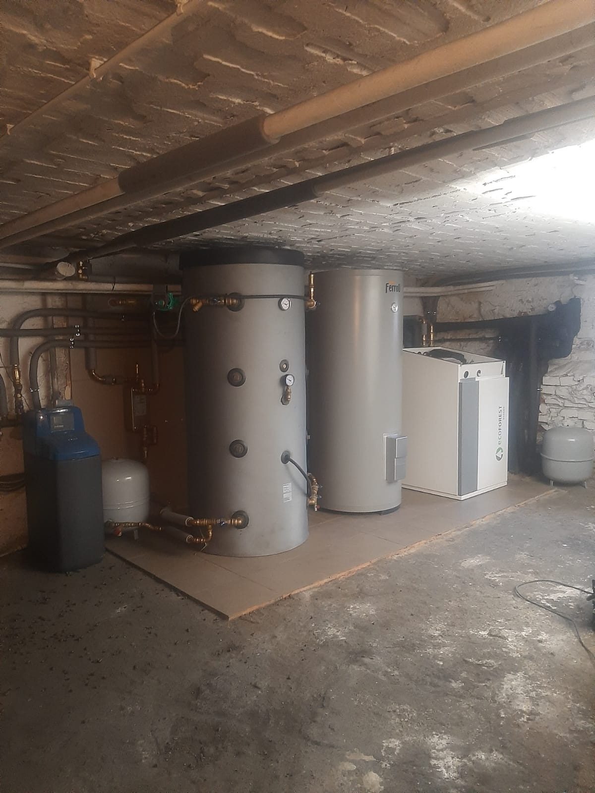 Kompletna instalacja grzewcza z dwoma zbiornikami Ferroli, pompą ciepła Ecoforest i zmiękczaczem wody w piwnicy z odsłoniętym sufitem.