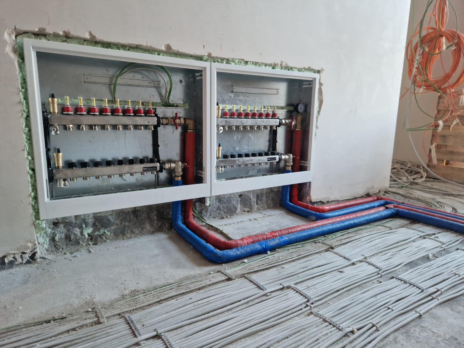 Rozdzielacz ogrzewania podłogowego w białej obudowie, z widocznymi rurami w izolacji czerwonej i niebieskiej, ułożonymi na szarym podłożu z instalacją. Etap prac w surowym wnętrzu.