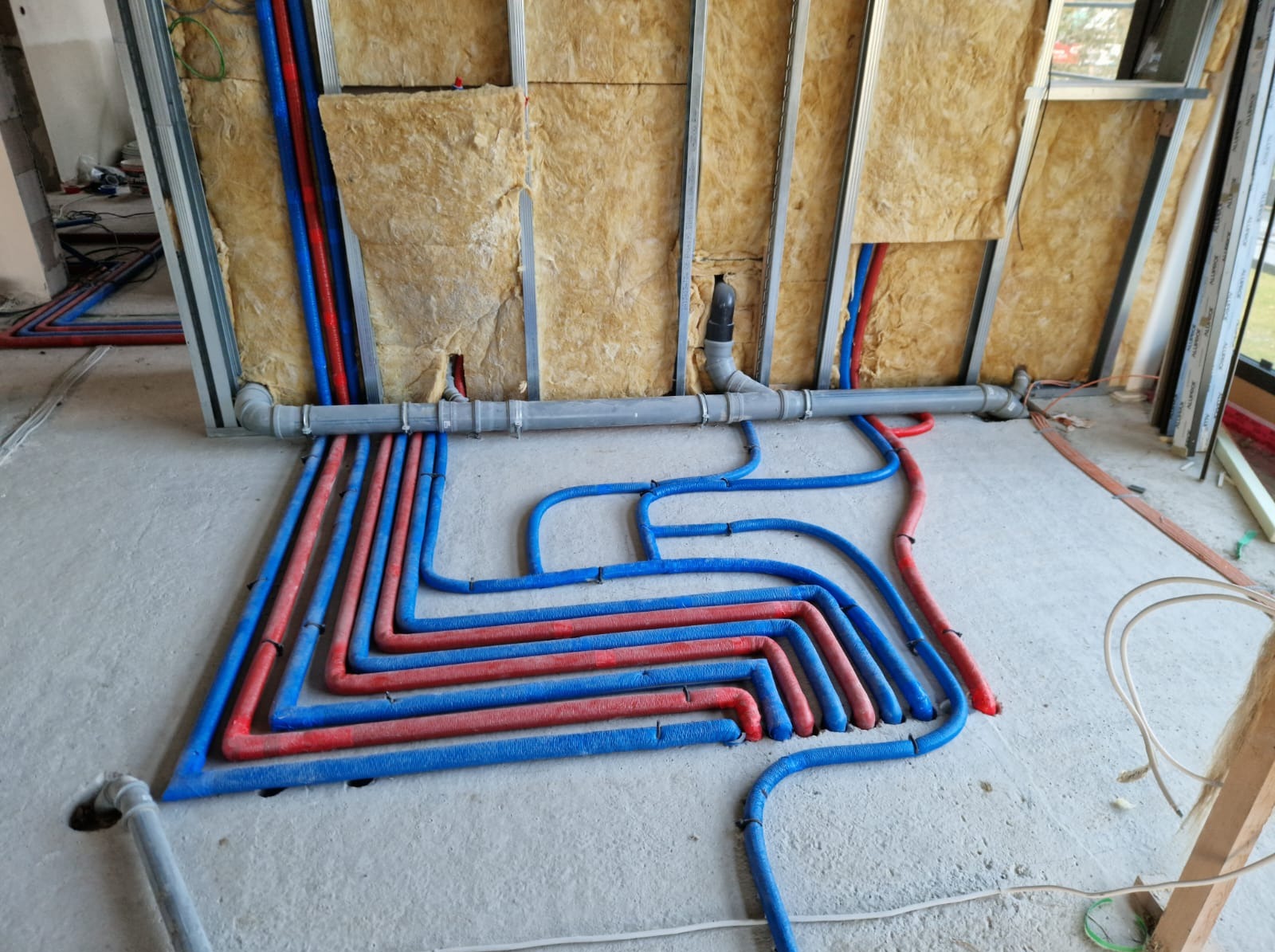Instalacja rur grzewczych (czerwono-niebieskie) w surowym wnętrzu, widoczne izolowane ściany z wełną mineralną i szare rury kanalizacyjne. Prace hydrauliczne w trakcie realizacji.