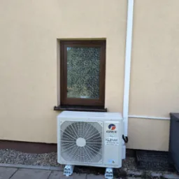 Zamontowana jednostka zewnętrzna klimatyzacji Gree Klimak na elewacji domu z brązowym oknem. Widoczna biała rura odprowadzająca skropliny i kostka brukowa.