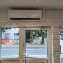 Klimak Wojciech Makowski - Biała klimatyzacja na ścianie z cegły nad przeszklonymi drzwiami wejściowymi. Widok na ulicę z zielenią i budynkami w tle.