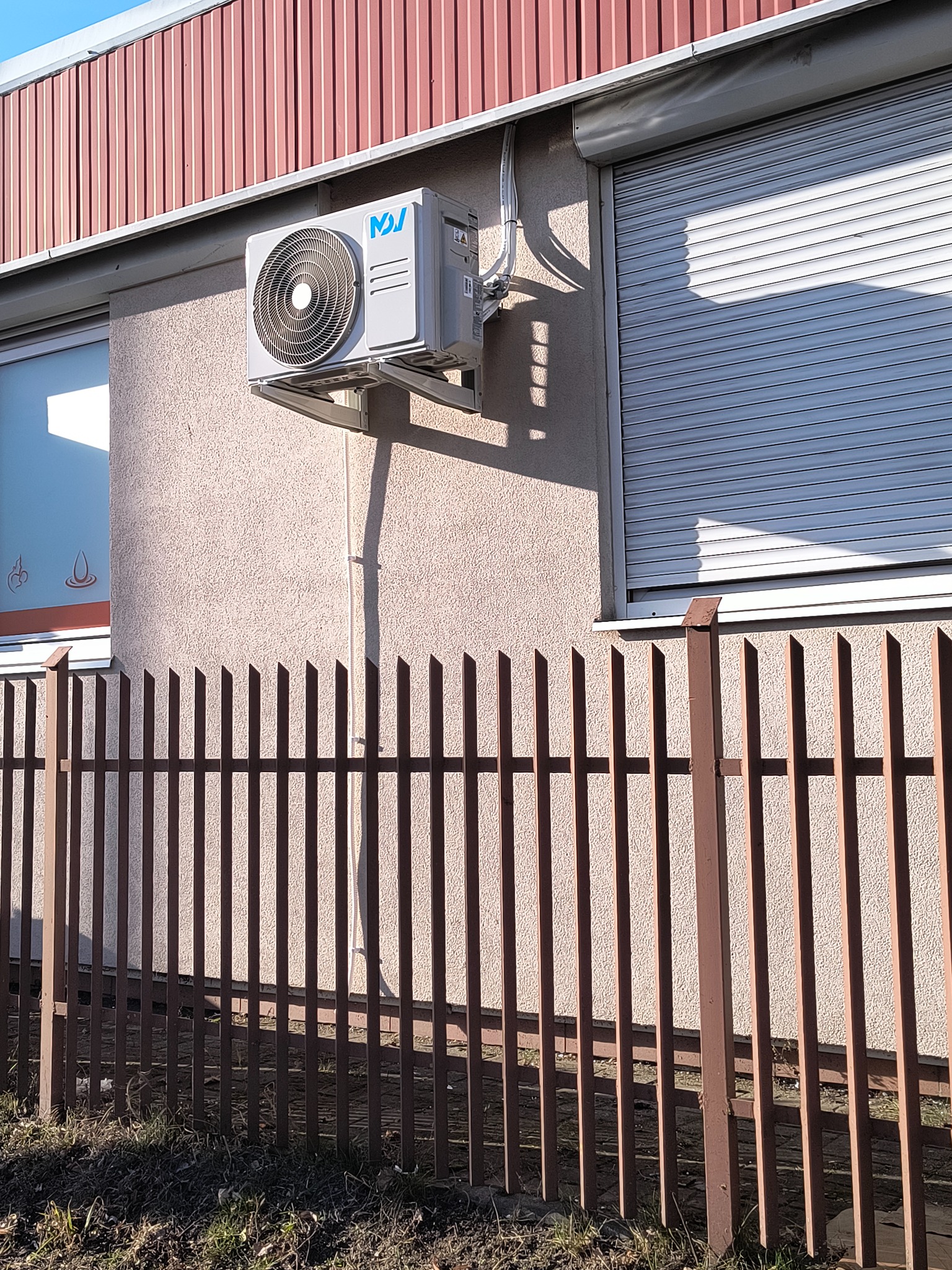 Zewnętrzny agregat klimatyzacji zamontowany na ścianie budynku obok zamkniętej rolety zewnętrznej, widoczny przez metalowy płot.