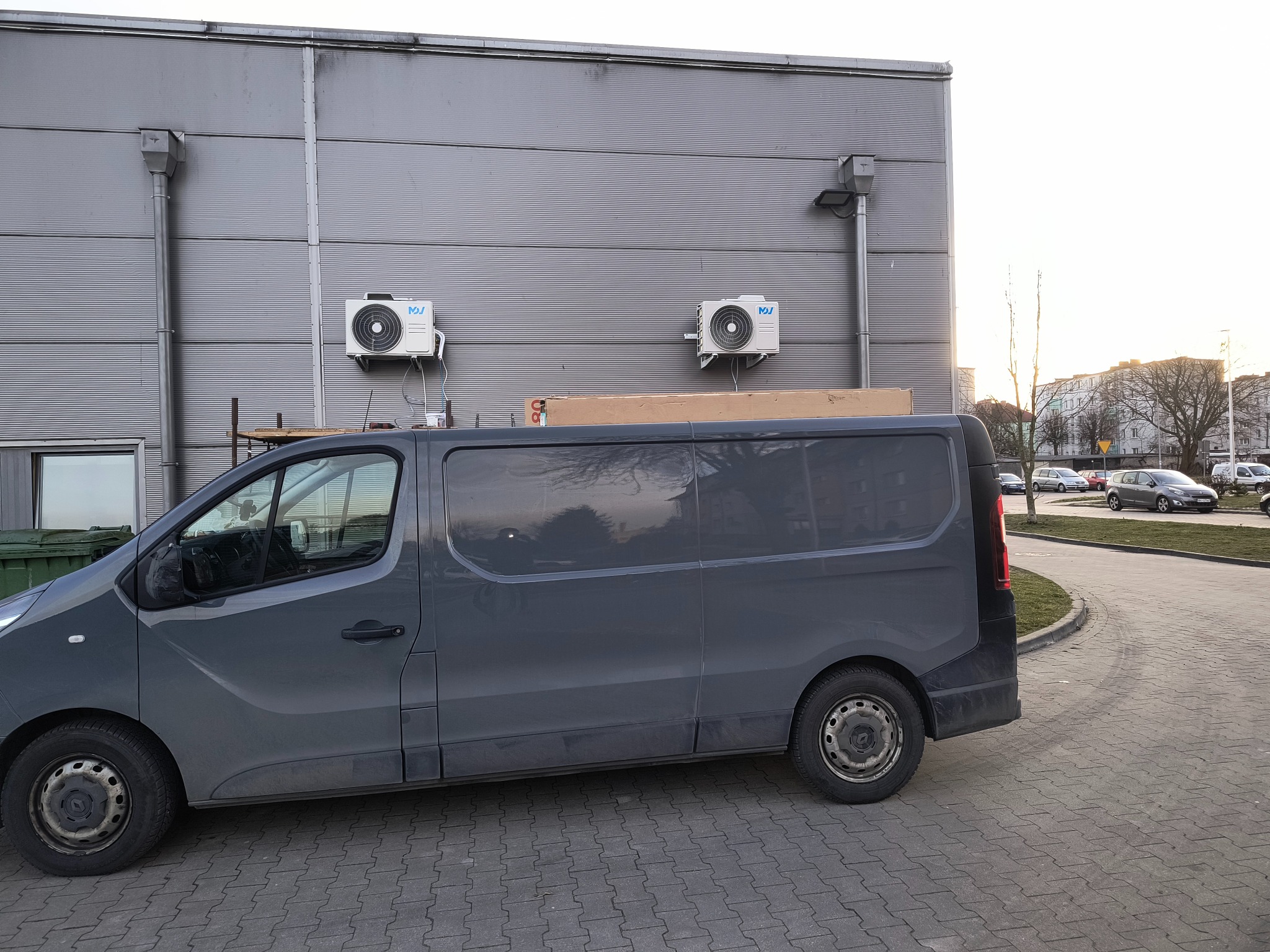 Dwa klimatyzatory zamontowane na szarej ścianie budynku przemysłowego, widoczne zza zaparkowanego szarego furgonetki dostawczej z kartonem na dachu.
