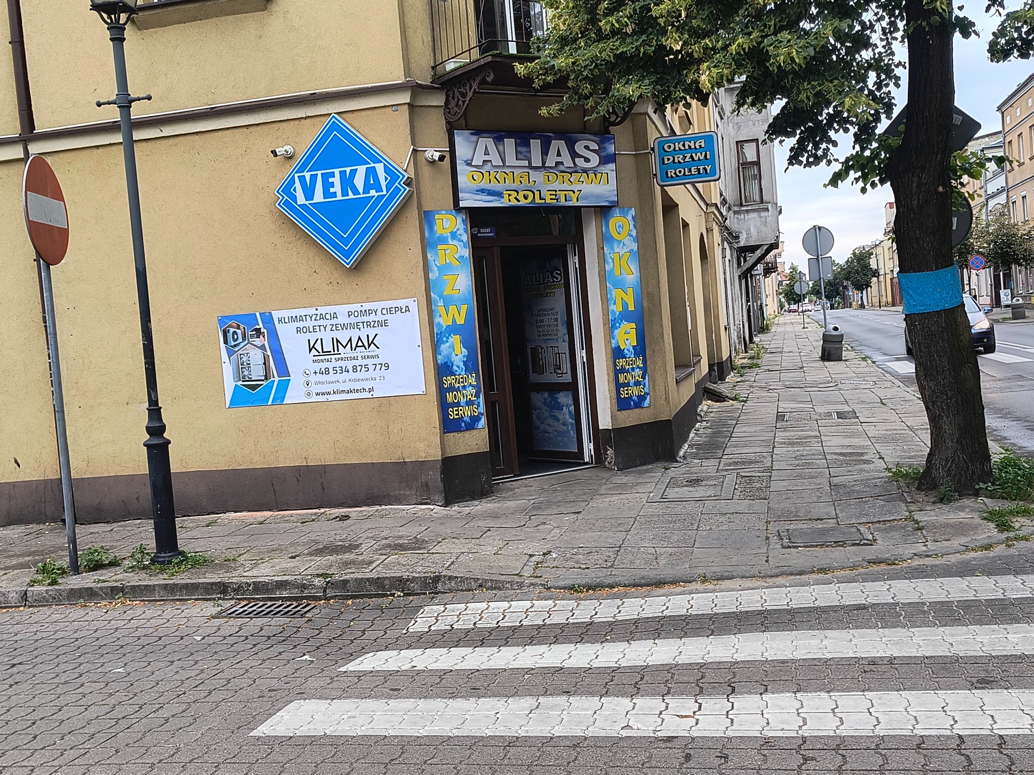 Fasada budynku z szyldem 'ALIAS OKNA, DRZWI, ROLETY' i reklamami Veka i Klimak. Widoczne drzwi wejściowe do sklepu i fragment chodnika z przejściem dla pieszych.